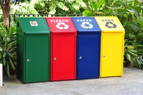 A row of four outdoor recycling bins positioned on a paved surface with lush green foliage and plants in the background. The bins are made of metal or durable plastic with a smooth, glossy finish and are arranged side by side, each designated for specific waste types. From left to right, the first is green with a label indicating general waste or non-recyclables, featuring a small white icon and no further text visible. The second bin is red, clearly marked for plastic recycling with a white recycling symbol and the word 'PLASTIC' in bold white letters. The third is blue, designated for paper recycling, with a white recycling icon and the word 'PAPER' prominently displayed. The fourth bin is yellow, intended for can recycling, with a recycling symbol and the word 'CANS' in bold black lettering. Each bin has a lid with a small locking mechanism or handle visible on the front, and they appear to be in good condition. This setup is typical for a commercial or residential area in Blackheath, supporting local waste diversion efforts managed by Commercial Waste Removal Blackheath, situated in the postcode area of London. The overall scene emphasizes organized waste segregation suitable for outdoor recycling collection in a UK environment.