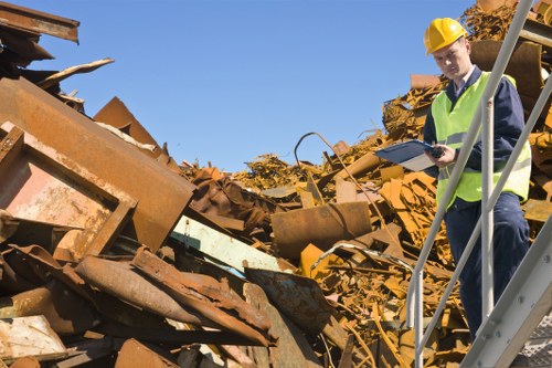 A construction site outside with a large pile of rusted metal debris, including sheets, beams, and scrap materials, scattered across the ground. The debris has a weathered, uneven surface with varying shades of reddish-brown and orange rust. In the background, a clear blue sky is visible, indicating good weather. A worker, wearing a yellow safety helmet and high-visibility vest, is standing on a metal staircase adjacent to the pile, holding a clipboard and inspecting the site. The environment suggests a waste management or rubbish clearance operation near Blackheath, with the focus on safely assessing and documenting scrap metal. The scene captures a professional approach to disposing of or managing construction and metal waste as part of commercial rubbish removal services provided by companies like Commercial Waste Removal Blackheath.