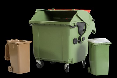 The image displays three wheelie bins arranged on a flat surface, likely an outdoor driveway or pavement. The central bin is the largest, with a green, high-density polyethylene body featuring a textured, slightly matte surface, and a matching green lid that is closed. This bin also has a black carry handle on the side and black wheel hubs, indicating mobility. To the left is a smaller, tan-colored wheelie bin with a similarly textured plastic finish and a closed lid, positioned slightly behind the larger bin. On the right is another small green bin, comparable in size to the tan one, also with a textured plastic surface and a closed lid, placed in front of the larger bin. The background is dark and nondescript, highlighting the bins in the foreground. This setup appears typical for local rubbish collection or waste management services, such as those offered by Commercial Waste Removal Blackheath, with the bins suitable for general waste or recyclable materials in a residential or commercial setting within or near Blackheath postcode area, South London.