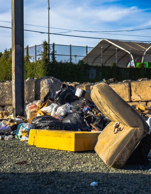 A worker wearing a green uniform and safety helmet is standing at the open back of a commercial rubbish collection van, which is parked on a residential street lined with trees and parked cars. The va