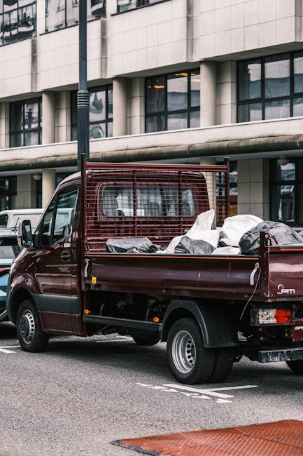 A small maroon flatbed truck parked on an urban street with a modern office building in the background. The truck's bed is filled with mixed waste materials, including large white and black plastic ba